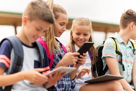 primary education, friendship, childhood, technology and people concept - group of happy elementary school students with backpacks sitting on bench and talking outdoorsの写真素材