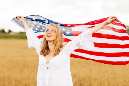 country, patriotism, independence day and people concept - happy smiling young woman in white dress with national american flag on cereal fieldの写真素材