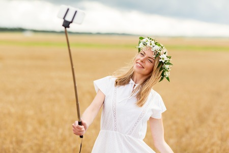 technology, summer holidays, vacation and people concept - smiling young woman in wreath of flowers taking picture by smartphone selfie stick on cereal fieldの写真素材