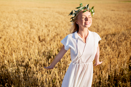 happiness, nature, summer holidays, vacation and people concept - smiling young woman in wreath of flowers and white dress on cereal fieldの写真素材