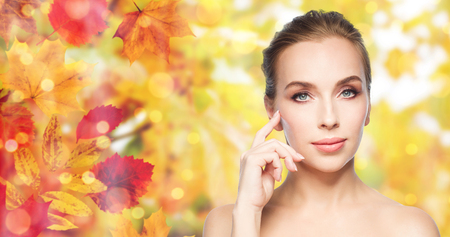 beauty, people and skin care concept - beautiful young woman showing her cheekbone over natural autumn leaves and lights backgroundの写真素材