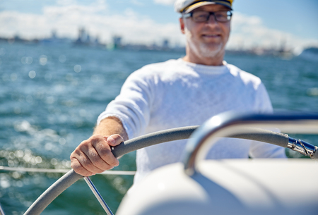 sailing, age, tourism, travel and people concept - close up of happy senior man in captain hat steering wheel and navigating sail boat or yacht floating in seaの写真素材