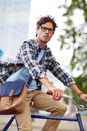people, style, leisure and lifestyle - young hipster man with shoulder bag and earphones riding fixed gear bike on city streetの写真素材