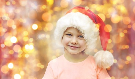 holidays, christmas, childhood and people concept - smiling little girl in santa hat sitting on couch over lights backgroundの写真素材