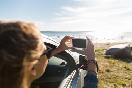 summer vacation, holidays, travel, road trip and people concept - happy teenage girl or young woman in car taking picture of sea with smartphoneの写真素材
