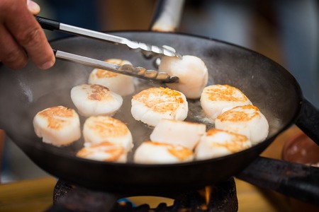 cooking, asian kitchen, sale and sea food concept - close up of cook with tongs frying scallops in cast iron pan at asian street marketの写真素材