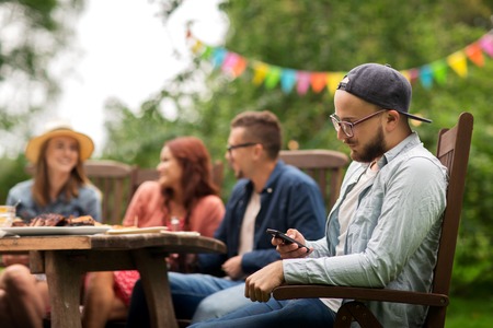 leisure, holidays, people and technology concept - young man texting on smartphone and friends having dinner at summer garden partyの写真素材