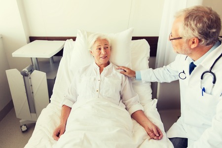 medicine, age, support, health care and people concept - doctor visiting and cheering senior woman lying in bed at hospital wardの写真素材