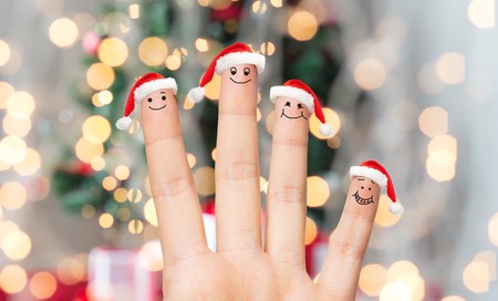family, holidays, christmas and body parts concept - close up of hand with four fingers in santa hats with smiley faces over lights backgroundの写真素材