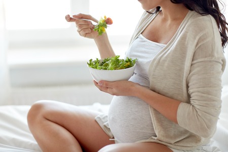 pregnancy, healthy food and people concept - close up of happy pregnant woman eating vegetable salad for breakfast in bed at homeの写真素材
