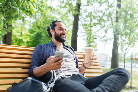 people,  technology, travel and tourism - man with earphones, smartphone and bag sitting on city street bench and listening to musicの写真素材