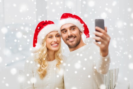christmas, holidays, technology and people concept - happy couple in santa hats taking selfie picture with smartphone at homeの写真素材