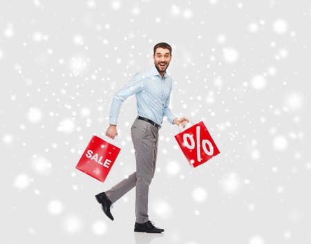 people, sale, christmas, winter and holidays concept - smiling man holding red shopping bags with percentage sign over snow backgroundの写真素材