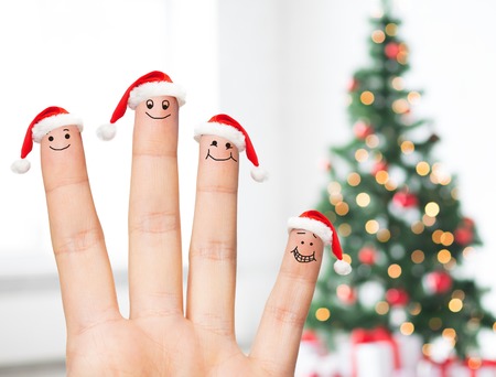 family, holidays, christmas and body parts concept - close up of hand with four fingers in santa hats with smiley faces over tree lights backgroundの写真素材