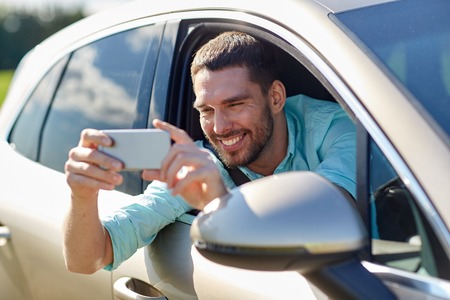 road trip, transport, travel, technology and people concept - happy smiling man with smartphone driving in car and taking selfieの写真素材
