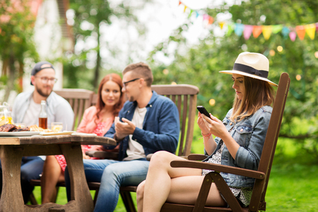 leisure, holidays, people and technology concept - young woman or teenage girl texting on smartphone and friends having dinner at summer garden partyの写真素材