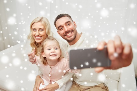 holidays, technology and people concept - happy family sitting on sofa and taking selfie picture with smartphone at homeの写真素材