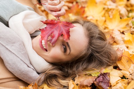 season and people concept - beautiful young woman with autumn maple leaf lying on groundの写真素材