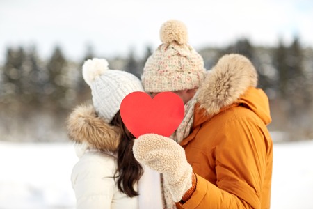 people, season, love, christmas and valentines day concept - happy couple hiding faces behind blank red heart over winter landscapeの写真素材
