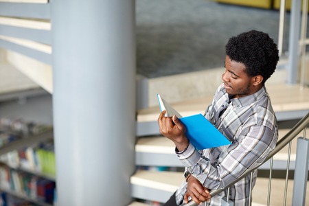 education, high school, university, learning and people concept - happy african american student boy or young man reading book on stairs at libraryの写真素材