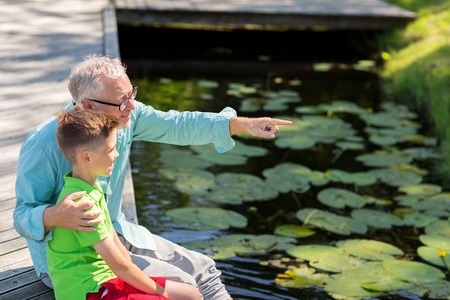 family, generation, communication and people concept - happy grandfather and grandson sitting on river berthの写真素材