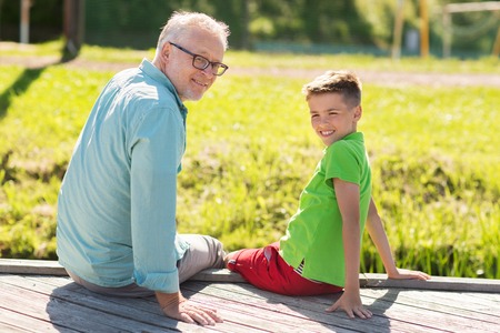 family, generation, communication and people concept - happy grandfather and grandson sitting on river berthの写真素材