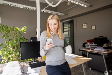pregnancy, business, work and technology concept - smiling pregnant businesswoman with smartphone at office tableの写真素材