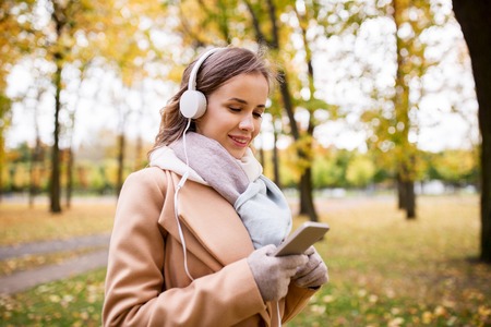 season, technology and people concept - beautiful happy young woman with headphones listening to music on smartphone walking in autumn parkの写真素材