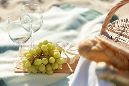 food, holidays, celebration and summer concept - close up of picnic basket with grapes, wine glasses and cheese on beachの写真素材
