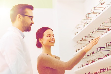health care, people, eyesight and vision concept - happy woman choosing and showing glasses to optician at optics storeの写真素材