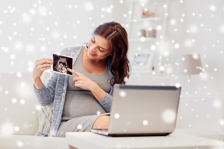 pregnancy, motherhood, people and expectation concept - happy pregnant woman with laptop computer having video call and showing ultrasound image at home over snowの写真素材