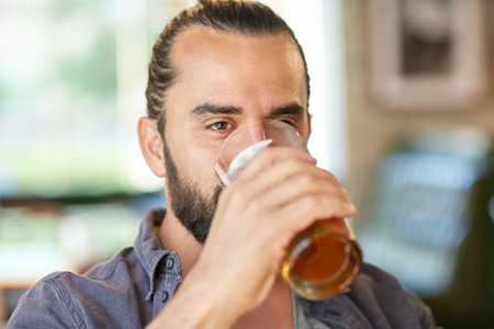 people, drinks, alcohol and leisure concept - close up of young man drinking beer from glass at bar...