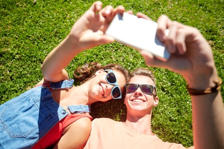 love and people concept - happy teenage couple in sunglasses lying on grass and taking selfie on...