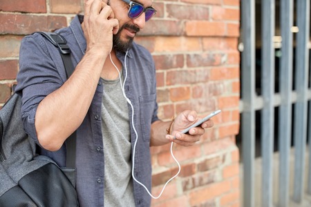 people, technology, travel and tourism concept - close up of man with earphones, smartphone and bag...