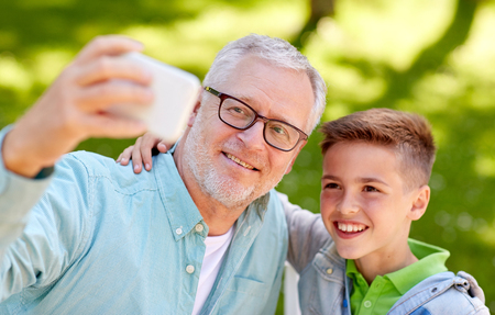 family, generation, technology and people concept - happy grandfather and grandson with smartphone taking selfie at summer parkの写真素材