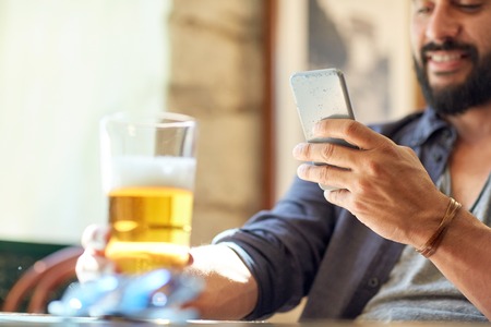 people, leisure and technology concept - close up of man with smartphone drinking beer and reading message at bar or pubの写真素材