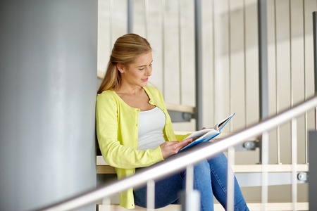education, high school, university, learning and people concept - smiling young woman or student girl reading book sitting on stairsの写真素材