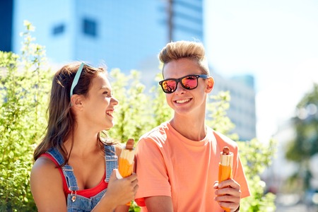 fast food, summer and people concept - happy teenage couple eating hot dogs sitting on city street benchの写真素材