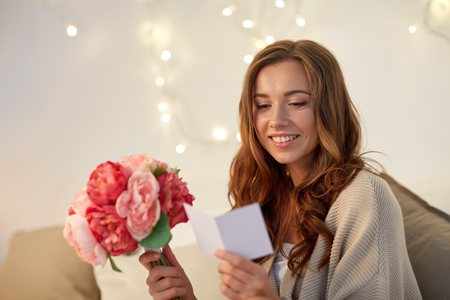 holidays, winter, birthday and people concept - happy young woman with flowers reading greeting card in bed at home bedroomの写真素材