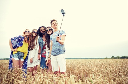nature, summer, youth culture, technology and people concept - smiling young hippie friends in sunglasses taking picture by smartphone on selfie stick on cereal fieldの写真素材