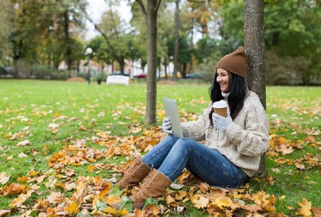 season, technology and people concept - young woman with tablet pc and coffee cup sitting on grass in autumn parkの写真素材