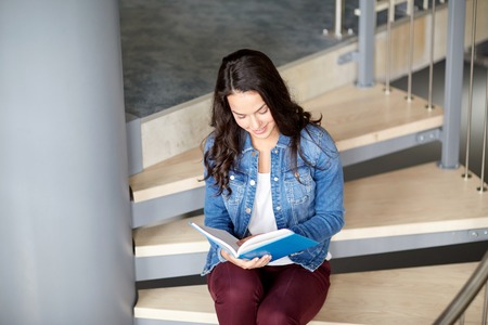 education, high school, university, learning and people concept - smiling student girl reading book sitting on stairsの写真素材