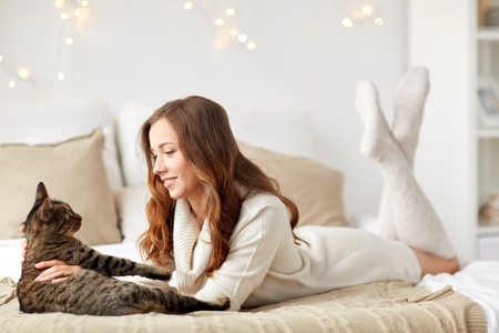 pets, comfort, christmas, winter and people concept - happy young woman with cat lying in bed at homeの写真素材