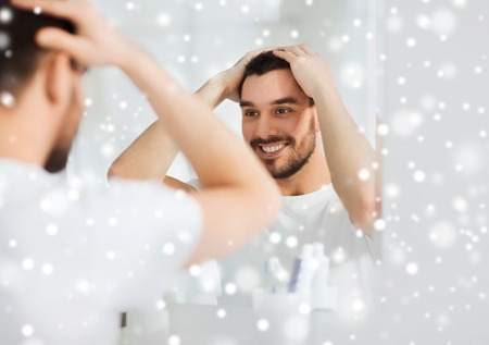 beauty, hygiene, hairstyle and people concept - smiling young man looking to mirror and styling hair at home bathroom over snowの写真素材