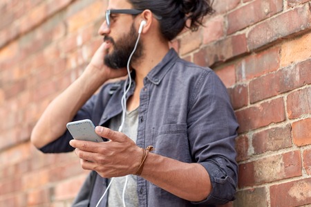people and technology concept - close up of man with earphones and smartphone listening to music at brick wall on streetの写真素材