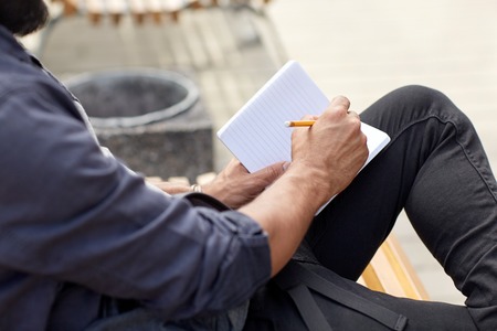 lifestyle, freelance, inspiration and people concept - close up of man with bag writing to notebook or diary sitting on city street benchの写真素材