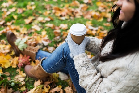 season, technology and people concept - close up of happy young woman drinking coffee from paper cup in autumn parkの写真素材