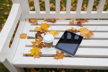 season, technology and advertisement concept - tablet pc computer and coffee paper cup on bench in autumn parkの写真素材