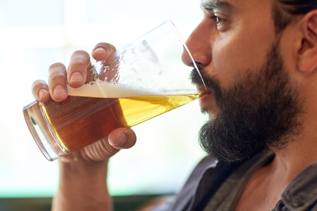 people, drinks, alcohol and leisure concept - close up of young man drinking beer from glass at bar or pubの写真素材