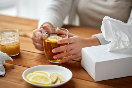 health, traditional medicine and ethnoscience concept - close up of ill woman drinking tea with lemon, honey and ginger at wooden tableの写真素材